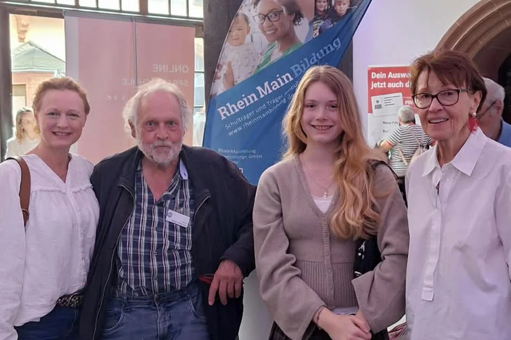 Vier Personen unterschiedlichen Alters (drei Frauen, ein älterer Mann) lächeln stehend vor einem Informationsstand mit Banner RheinMain Bildung in einem hellen Innenraum.