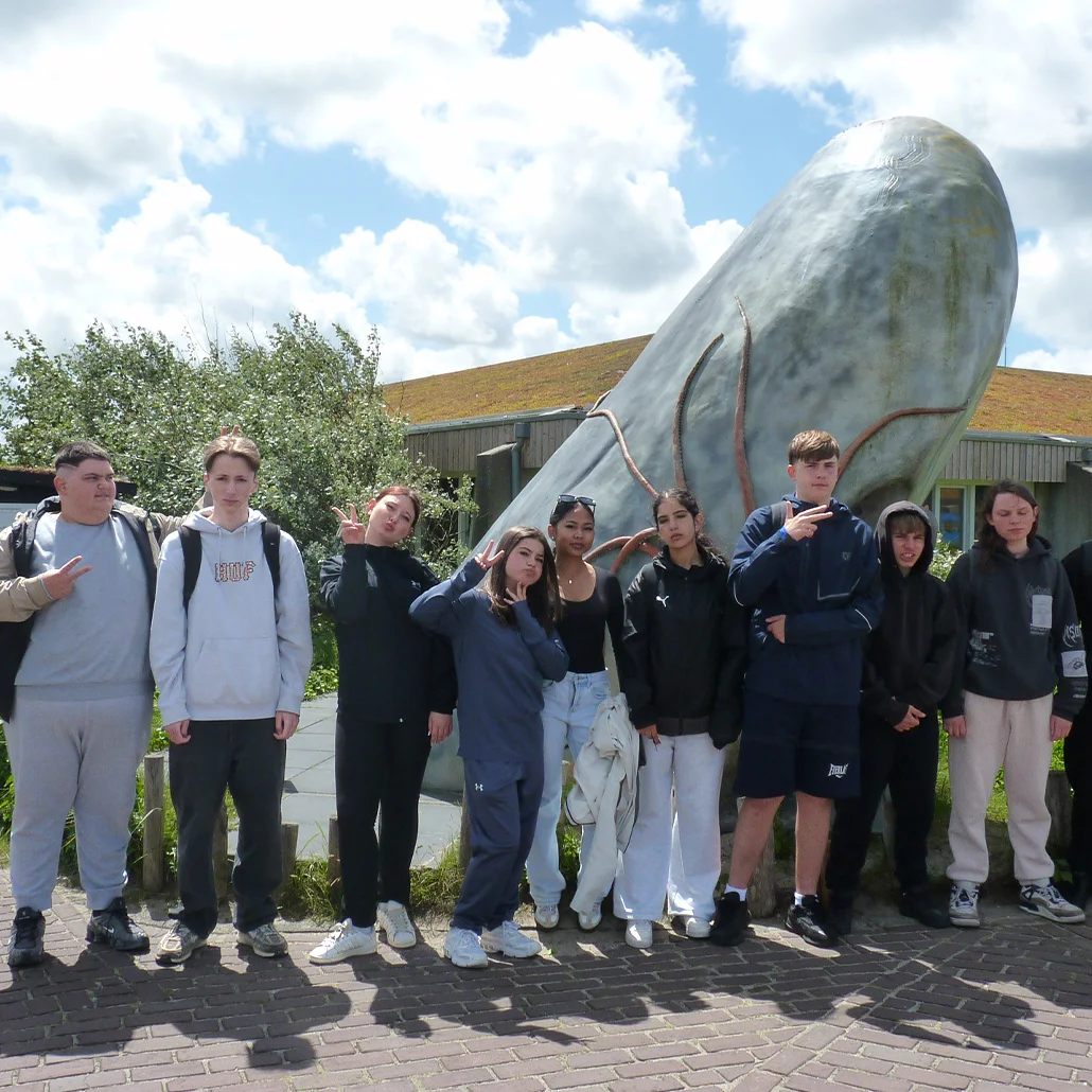 Gruppe Jugendlicher steht nebeneinander vor einer großen silbernen Walskulptur im Freien.