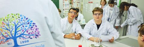 Das Bild zeigt im Vordergrund zwei Jugendliche an einem Tisch, im Hintergrund weitere Jugendliche