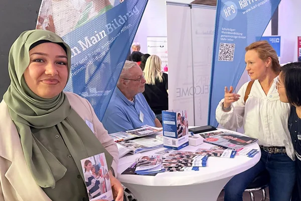 Diverse Messestand-Szene: Junge Frau mit Kopftuch lächelt und hält einen Flyer vor einem Stehtisch mit Broschüren; zwei Frauen unterhalten sich mit einem älteren Mann, Informationsbanner im Hintergrund.